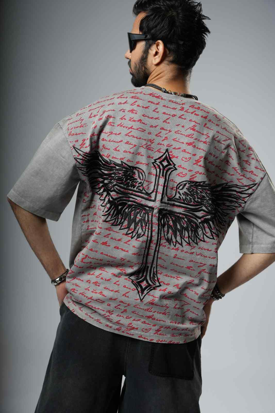Person wearing a t-shirt with a cross and wings design on the back, standing against a gray background.